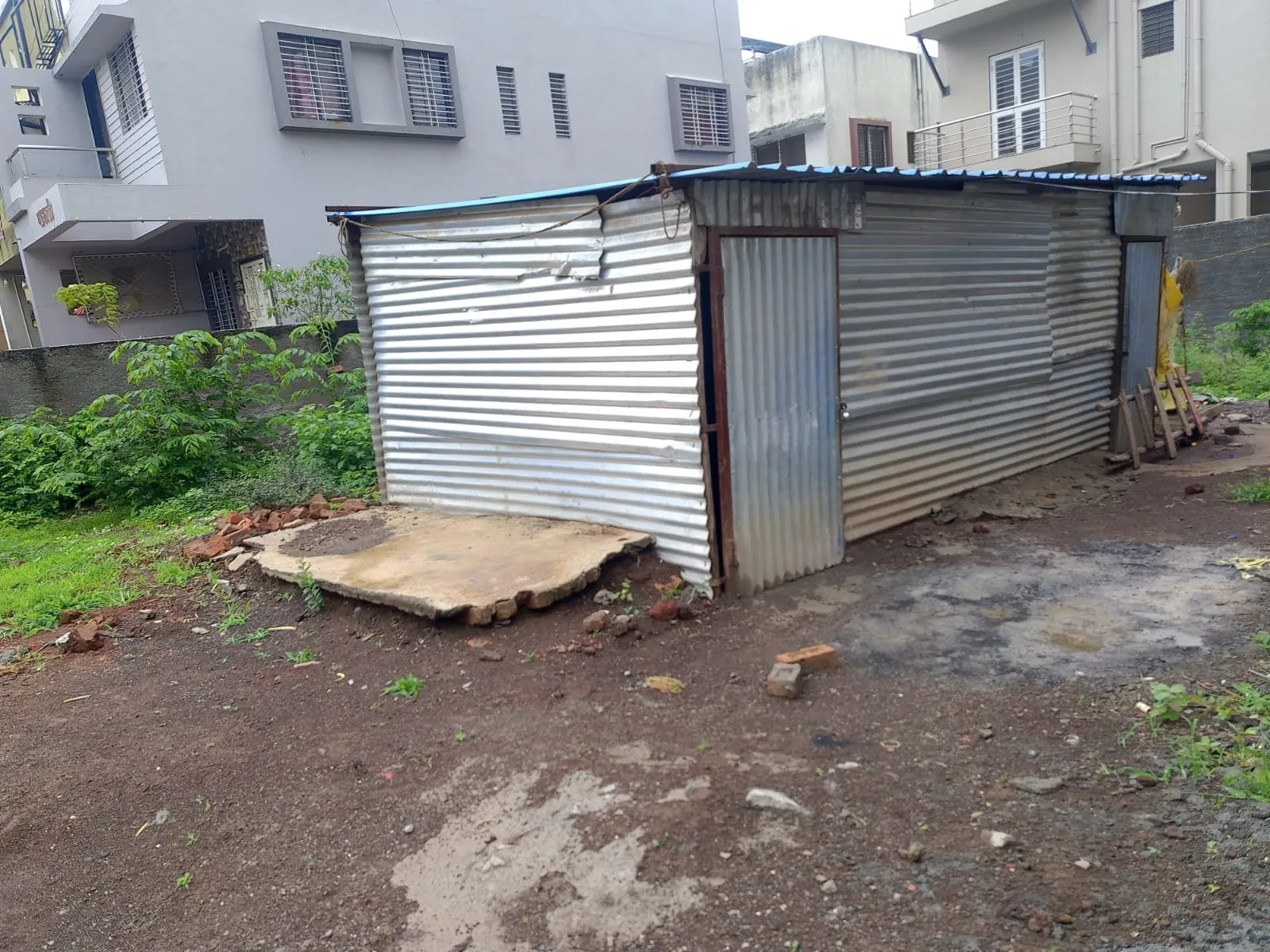 On-site view of the Adgaon plot with neighbouring homes and a temporary shed on the land